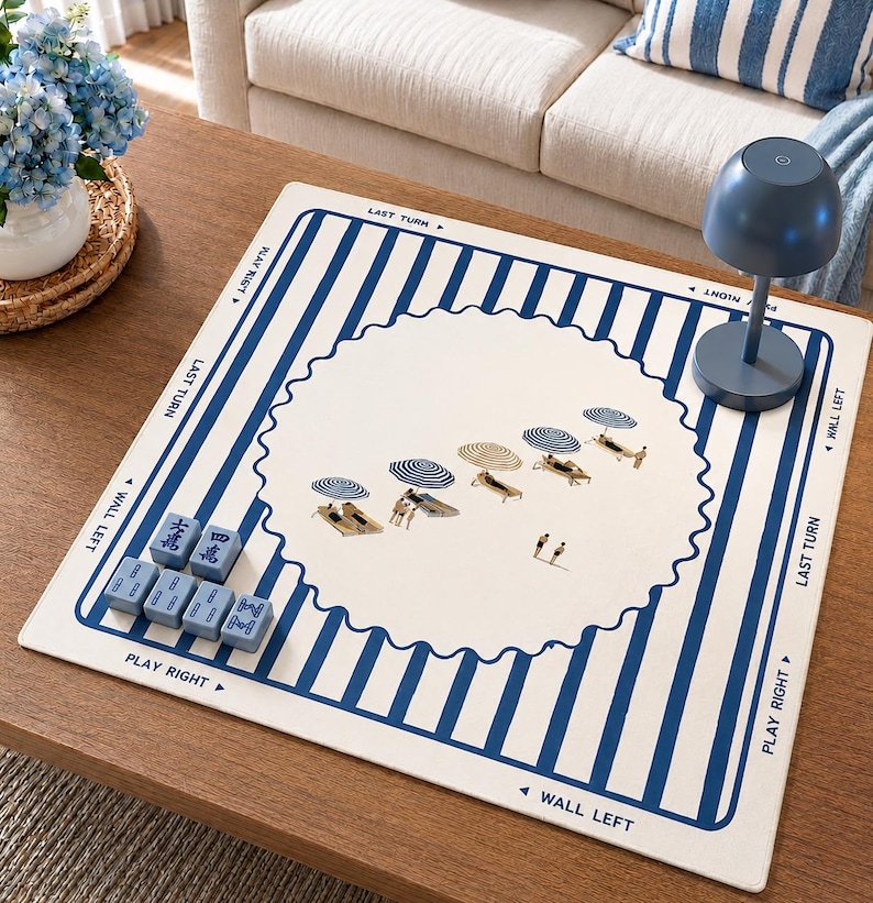 Puede incluir: Una alfombrilla de juego cuadrada con un centro blanco de borde festoneado, decorada con peque&ntilde;as ilustraciones de sombrillas de playa y tumbonas. El borde tiene rayas verticales azules y blancas con texto que incluye LAST TURN, PLAY RIGHT y WALL LEFT.