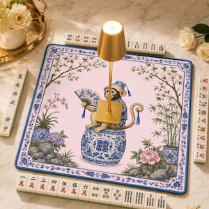 May include: A square mahjong mat with a light pink center featuring a monkey sitting on a blue and white porcelain stool, holding a folding fan. The mat has a blue and white floral border with bamboo and flower illustrations. A gold lamp sits on top.