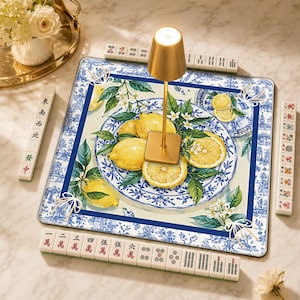 May include: A square mahjong mat featuring a blue and white chinoiserie pattern with yellow lemons and green leaves. A gold-toned cordless lamp sits in the center. The mat is surrounded by ivory-colored mahjong tiles with red and black markings.