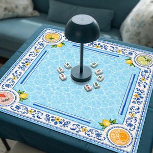 Peut inclure: Un tapis de mah-jong carré avec une surface bleu clair à motif d'eau, bordé de motifs floraux bleus et jaunes et d'illustrations d'agrumes. Une lampe de table gris foncé en forme de champignon est posée au centre avec des tuiles.