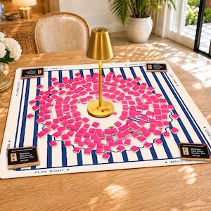 May include: A square Mah Jongg play mat featuring a white background with blue vertical stripes, marked with "PLAY RIGHT" and "WALL LEFT" text. A gold-colored lamp sits in the center, surrounded by numerous small, bright pink square tiles.