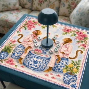 May include: A dark blue mushroom-shaped table lamp sits on a square fabric mat featuring an illustration of two monkeys in pink striped pajamas sitting on blue and white ceramic stools, surrounded by mahjong tiles and pink floral accents.