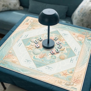 Op de afbeelding: Een vierkante Mahjong-mat met een lichtblauw en beige kustthema, versierd met illustraties van schelpen, zeesterren en koraal. De mat bevat gedrukte spelinstructies en een donkergrijze paddenstoelvormige lamp in het midden.
