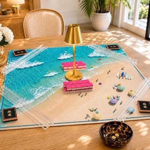 May include: A rectangular mahjong mat featuring a beach and ocean scene with turquoise water and white foam waves. The mat includes four clear acrylic tile racks, two black tiles with gold text, a gold lamp, pink tiles, and a leopard print bowl with dice.