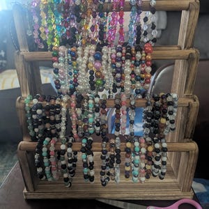 May include: A wooden display stand holding numerous beaded bracelets in various colours. The bracelets include clear, black, white, pink, and green beads. The stand has three tiers, each displaying multiple bracelets. Pink scissors are on the table.