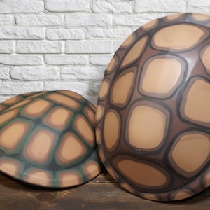 May include: Two turtle shell props. The shells are painted with a brown, tan, and green pattern. The larger shell is in the foreground, and the smaller shell is in the background. The shells are on a wooden surface.