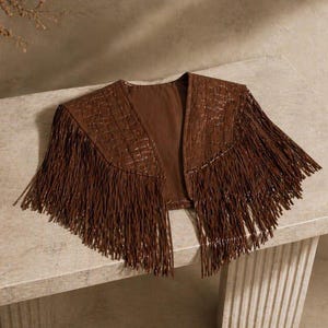 Puede incluir: Una chaqueta bolero corta de color marrón con un patrón texturizado de relieve de cocodrilo y largos flecos fluidos a lo largo del dobladillo y las mangas. La prenda tiene un diseño frontal abierto, forro interior liso y hombros estructurados.