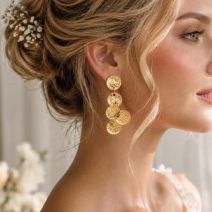 May include: A pair of gold-toned drop earrings featuring four circular discs with a textured, hammered finish. The discs are arranged in a cascading vertical line, suspended from a single stud attachment that secures to the earlobe.