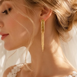 May include: A long, gold-toned dangle earring featuring a slender, rectangular frame encrusted with small, clear crystals. The lower portion of the earring consists of multiple delicate, fine gold chains that hang vertically, creating a fringe effect.