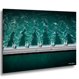Op de afbeelding: Luchtfoto van een brug over woelig turquoise water. De brug heeft een weg met auto's en een reeks structuren. Het wateroppervlak vertoont witte, schuimige patronen. De afbeelding is omlijst met een zwarte rand.