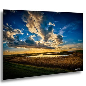 Op de afbeelding: Landschapsfoto van een zonsondergang boven een rivier. De lucht is een mix van blauw en goud, met dramatische wolken. De voorgrond toont hoog, goudkleurig riet en groene velden. Het beeld is ingelijst en klaar om op te hangen.