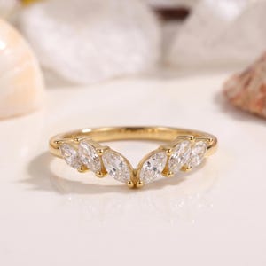 May include: A gold ring with a curved design. The ring features marquise-cut, clear gemstones set in a symmetrical pattern. The ring is displayed on a white surface. The gemstones are set in a delicate band.