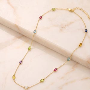 May include: A gold necklace with a delicate chain, adorned with a series of oval-shaped gemstones in various colours. The gemstones include red, blue, green, yellow, and purple. The necklace is presented on a light surface.