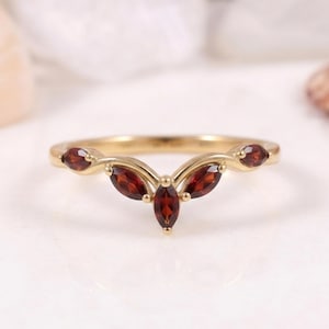 May include: A gold chevron-shaped ring featuring five marquise-cut deep red garnet gemstones set in a row. The band has a polished finish and follows a V-shaped contour, with the central garnet being the largest and flanked by four smaller stones.