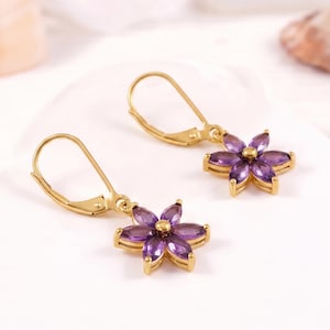 May include: A pair of gold-toned leverback earrings featuring a flower-shaped design. Each earring has six marquise-cut purple amethyst-coloured stones arranged as petals around a central gold-toned bead, creating a floral aesthetic.