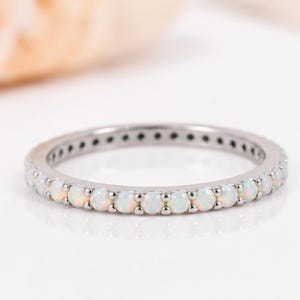 Könnte beinhalten: Ein dünner, kreisförmiger Ring aus poliertem, silberfarbenem Metall mit einer durchgehenden Reihe kleiner, runder, irisierender weißer Opal-Edelsteine, die gleichmäßig in einer Krappenfassung um den gesamten Ringumfang angeordnet sind.