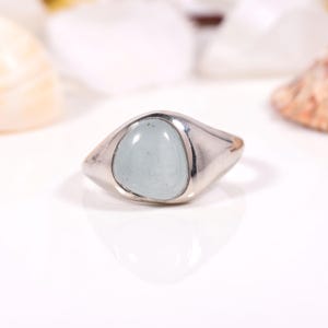 Könnte beinhalten: Ein silberner Ring mit einem hellblauen, oval geformten Stein. Der Ring hat eine glatte, polierte Oberfläche und ein klassisches Design. Der Stein ist in einer Lünettenfassung gefasst, und das Band verjüngt sich leicht.