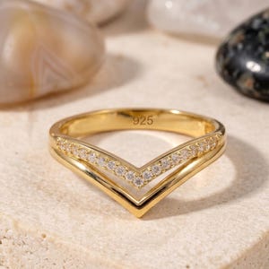 May include: A gold-coloured ring with a V-shaped design. The ring has a row of small, clear gemstones along the top V-shape. The number "925" is engraved on the inside. The ring is set against a neutral background.