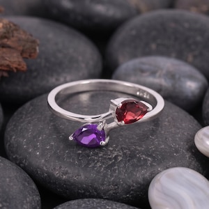 Op de afbeelding: Een zilveren bypass-ring met twee peervormige edelstenen, één dieprood en één levendig paars, gezet in een gepolijste metalen band. Het ontwerp toont een gespleten ringband die buigt om elke druppelvormige steen stevig in een zetting te houden.