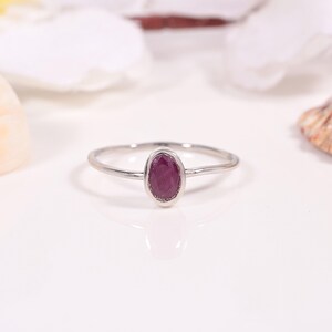 May include: A silver ring with a ruby gemstone. The oval-shaped ruby is a deep red color and set in a bezel setting. The ring has a thin band and is displayed on a white surface.