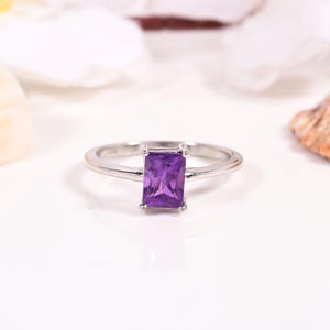 May include: A silver ring featuring a rectangular, deep purple gemstone. The ring has a thin, polished band, and the gemstone is set in a four-prong setting. The ring is presented on a white surface.