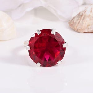 May include: A silver ring with a large, round, deep red gemstone. The gemstone is held by six silver prongs. The ring is set against a white background with a seashell and white fabric in the background.