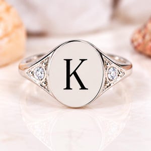 May include: A silver-toned signet ring featuring a smooth, polished oval face engraved with the letter K in a serif font. The ring band includes two small, clear, round-cut stones set into triangular metal accents on each side of the center.