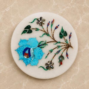 May include: A circular white marble coaster featuring an intricate inlay design of a large bright blue flower and a delicate vine with smaller dark gray flowers, green leaves, and thin stems accented with red and blue stone details.