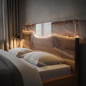 Puede incluir: Un cabecero de cama de madera con una parte superior de resina transparente e iluminada. El cabecero presenta un patrón de vetas de madera natural y un brillo cálido y ambiental. La cama está hecha con almohadas blancas y una manta gris.