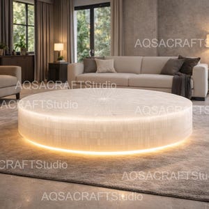 Op de afbeelding: Een moderne woonkamer met een grote, ronde, verlichte salontafel. De tafel heeft een zacht, gloeiend licht rond de basis. De kamer omvat een bank, vloerkleed en planten, waardoor een comfortabele en stijlvolle ruimte ontstaat.