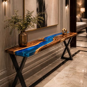Puede incluir: Una mesa consola larga que presenta una losa de madera natural con una incrustación de resina azul vibrante en forma de río en el centro. La mesa está sostenida por patas de metal negro en forma de X y sostiene un jarrón metálico con vegetación.