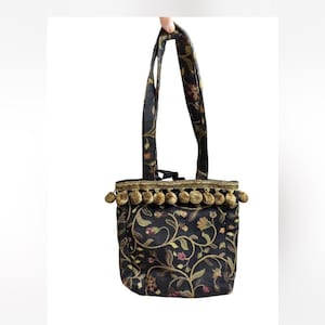 May include: A small black tote bag with a floral pattern in gold, green, and pink. The bag has a gold trim with small tassels and matching handles. The bag is held up by a hand.