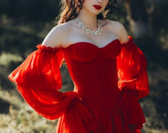 May include: A vibrant red off-the-shoulder gown with flowing sleeves and a layered skirt. The dress features a sweetheart neckline and is paired with a delicate silver necklace. The model has long, curly brown hair and red lipstick.