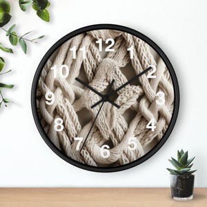 May include: A round wall clock with a black frame. The clock face features a beige, woven pattern with white numerals and black clock hands. The clock is on a wooden surface with a small potted plant.