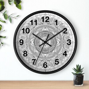 Op de afbeelding: Ronde zwarte wandklok met een gedetailleerd grijs en wit mandala-ontwerp. De wijzerplaat heeft zwarte cijfers en wijzers. De klok staat op een houten oppervlak met een kleine plant in een pot rechts.
