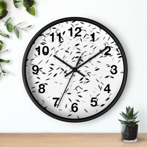 May include: A round wall clock with a black frame. The clock face is white with black numbers and a pattern of small, black leaf-like shapes. The clock hands are black.
