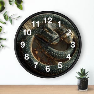 May include: Round black wall clock featuring a detailed image of a coiled snake. The snake is primarily dark green with gold accents, and the clock face has white numerals. The clock is set against a neutral background.