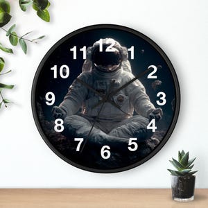 Pode incluir: Relógio de parede redondo preto com um astronauta em pose meditativa contra um fundo espacial escuro. O mostrador do relógio tem algarismos e ponteiros brancos, com uma moldura preta. Um item de decoração para casa com tema espacial.