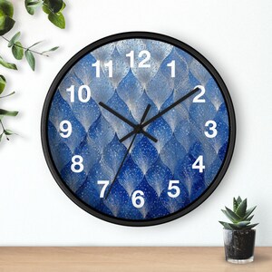 Könnte beinhalten: Runde Wanduhr mit schwarzem Rahmen und blau-weiß gemustertem Zifferblatt. Das Zifferblatt zeigt ein sich wiederholendes Design aus stilisierten Blättern. Weiße Ziffern markieren die Stunden, und schwarze Zeiger zeigen die Zeit an.