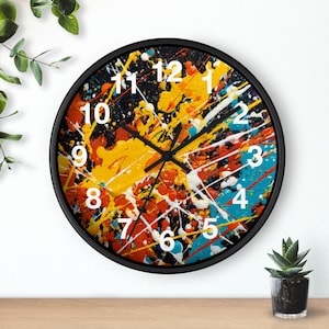 Puede incluir: Un reloj de pared redondo con marco negro. La esfera presenta un diseño abstracto con salpicaduras de pintura amarilla, naranja, blanca y azul. Los números blancos marcan las horas. El reloj está sobre una superficie de madera.