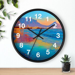 Puede incluir: Reloj de pared redondo con marco negro. La esfera del reloj presenta una colorida escena de playa con una montaña al fondo. Números blancos marcan las horas. Las manecillas del reloj son negras.