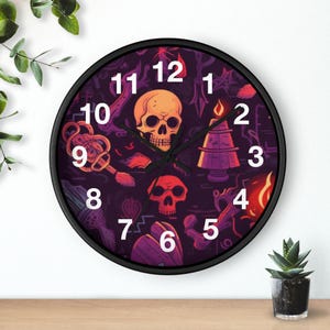 Op de afbeelding: Ronde zwarte wandklok met een griezelig ontwerp. De wijzerplaat toont een schedel, kaars en andere Halloween-thema graphics in oranje, paars en wit. De wijzers zijn zwart en de cijfers wit.
