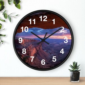 Könnte beinhalten: Runde Wanduhr mit schwarzem Rahmen. Das Zifferblatt zeigt eine malerische Canyonlandschaft bei Sonnenuntergang, mit weißen Ziffern und schwarzen Zeigern. Die Uhr steht auf einer Holzoberfläche.