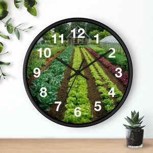 Könnte beinhalten: Eine runde Wanduhr mit schwarzem Rahmen. Das Zifferblatt zeigt ein lebendiges Bild eines Gemüsegartens mit Reihen von Feldfrüchten, einer kleinen Holzstruktur und einem Gewächshaus. Schwarze Zeiger zeigen auf die Zahlen auf dem Zifferblatt.