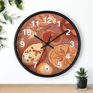 Könnte beinhalten: Eine runde Wanduhr mit schwarzem Rahmen. Das Zifferblatt hat einen braunen Hintergrund mit Illustrationen von Backwaren, Früchten und Blättern. Weiße Zahlen markieren die Stunden. Die Zeiger der Uhr sind schwarz.