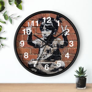 May include: Round wall clock with a black frame featuring a stencil art design on a brick background. The artwork depicts a child making a peace sign. White numbers mark the hours.