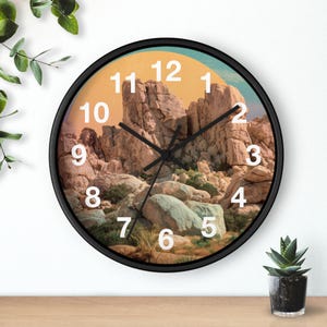 Könnte beinhalten: Runde Wanduhr mit schwarzem Rahmen, die eine Wüstenlandschaft zeigt. Das Zifferblatt zeigt eine Felsformation unter einem gelb-blauen Himmel, mit weißen Ziffern und schwarzen Zeigern. Eine kleine Topfpflanze steht auf einer Holzoberfläche.