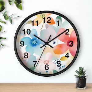 May include: A round wall clock with a black frame. The clock face features a watercolor floral design in shades of blue, pink, orange, and yellow. Black hands indicate the time. The clock is on a wooden surface.