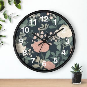 May include: Circular wall clock with a black frame and a floral pattern. The clock face has a dark blue backdrop with pink and cream flowers and green leaves. White numerals indicate the hours.