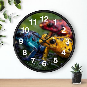 May include: A round wall clock with a black frame featuring a colorful image of three poison dart frogs. The frogs are blue, yellow, and red, with black spots. White numbers mark the hours against a green background.